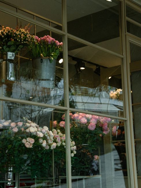 A display of fresh flowers visible through large glass windows of a flower shop. The arrangement includes pink tulips in a galvanized metal bucket, and clusters of pale pink and white roses and chrysa