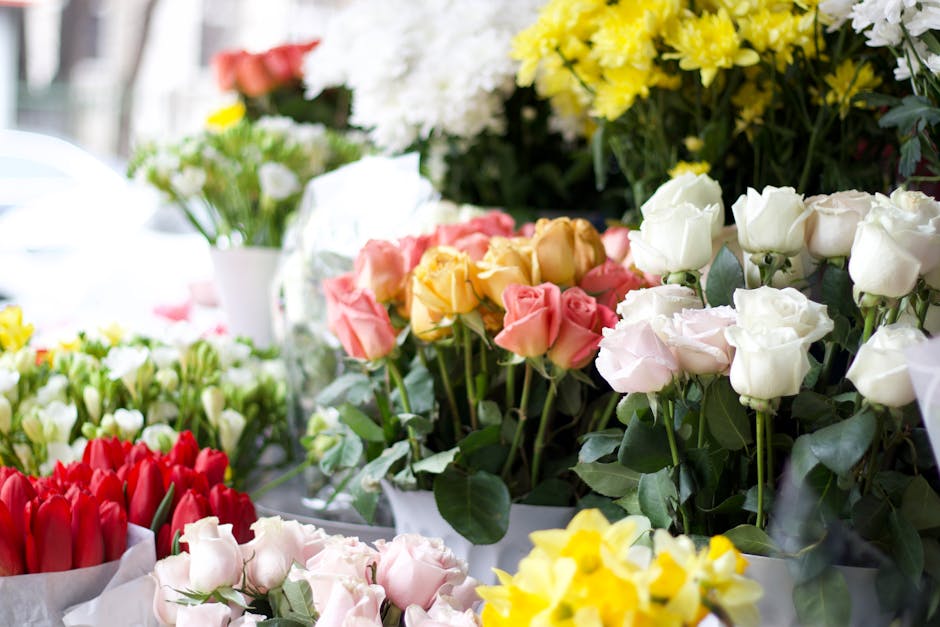A vibrant display of fresh flowers arranged in bouquets at a floral shop, featuring roses in shades of white, pale pink, coral, and bright yellow, alongside red tulips. The flowers are wrapped in whit