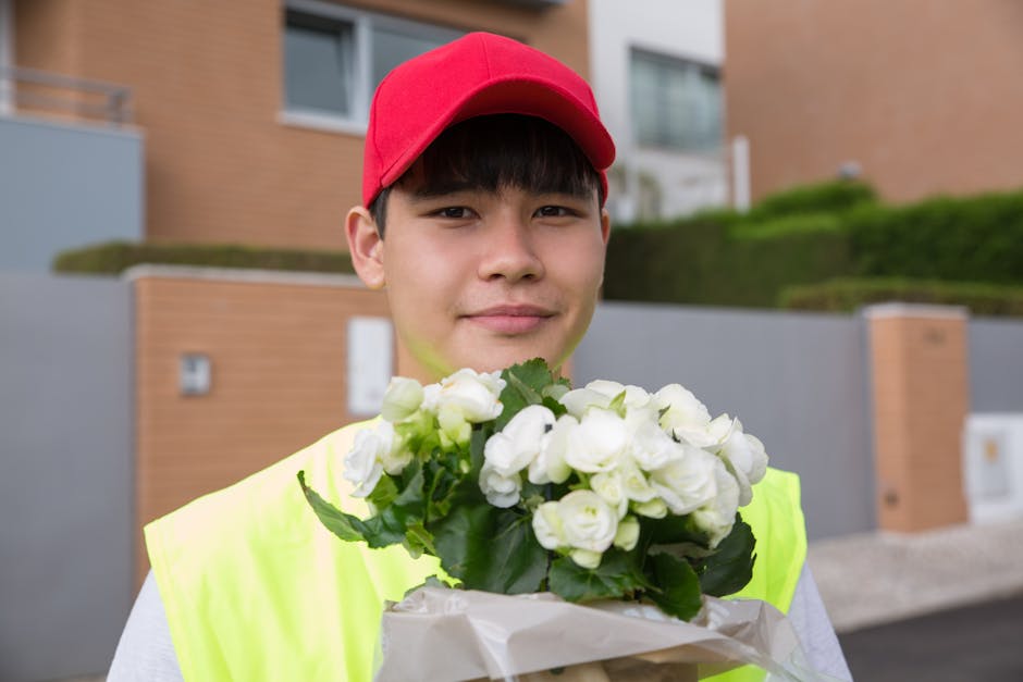 A young delivery person with fair skin, dark hair, and wearing a red cap and high-visibility yellow vest is holding a bouquet of fresh white roses and green foliage wrapped in beige paper. The arrange